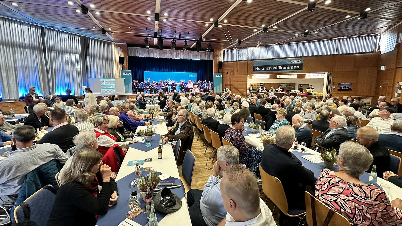 Gäste sitzen an langen Tischen im Bürgerhaus Froschhausen; auf der Bühne spielt ein Blasorchester.