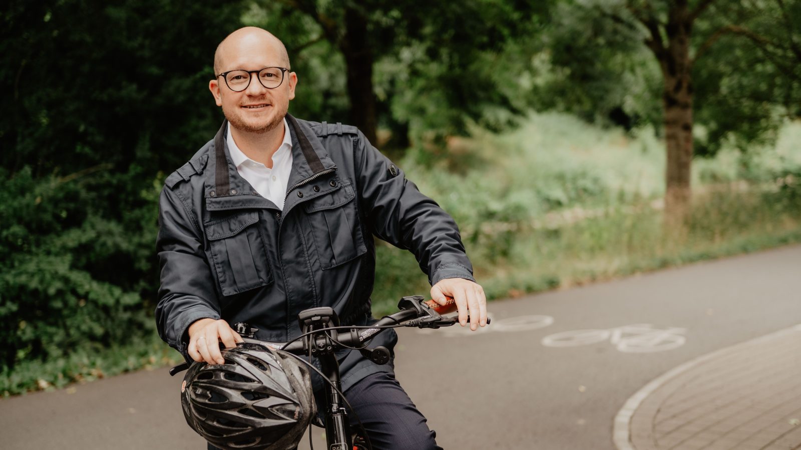 Rouven Kötter mit einem Fahrrad auf einem asphaltierten Radweg vor Bäumen.