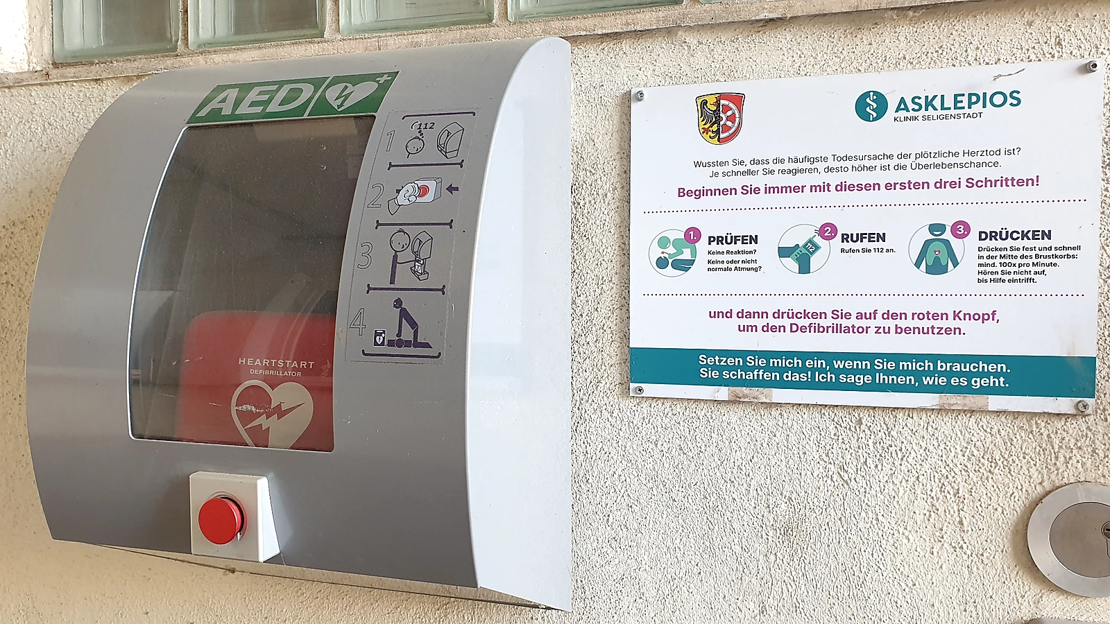 AED-Wandschrank mit Defibrillator, rotem Öffnungsknopf und Hinweistafel mit den Schritten Prüfen, Rufen, Drücken.